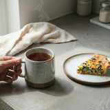 Person holding a mug of tea next to a slice of quiche on a kitchen counter.