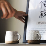 Two ceramic mugs on a surface with a blurred background and a framed text in the background.