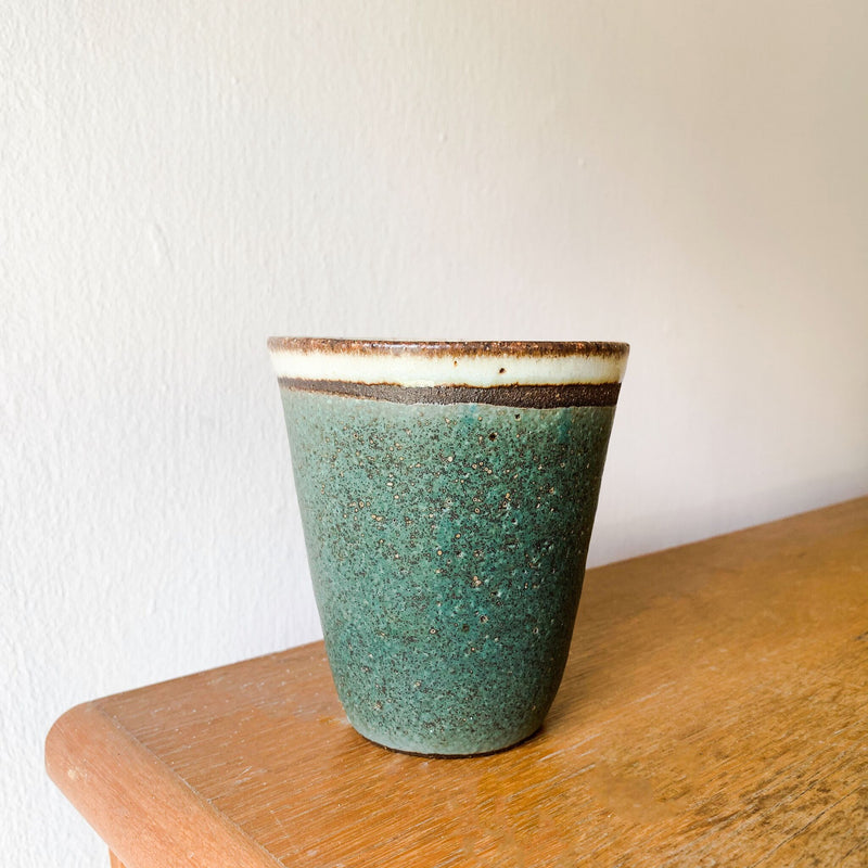 Matte green stoneware ceramic cup with a rustic and subtle white glaze at the rim, on top of a wooden platform.